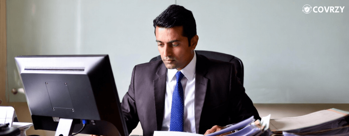 A man wearing a black suit working on his desk, a desktop computer and few documents lying infront of him