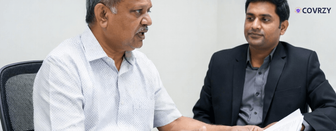 An Indian aged man wearing a white formal shirt talking with a man in a black suit. The aged man is holding a piece of document