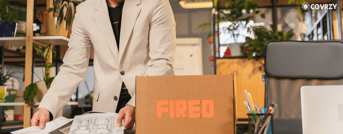 an employee in a formal suit with tucked in black t-shirt taking up some papers, beside lies a box labelled Fired