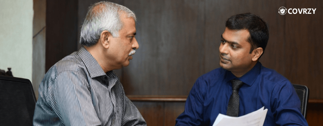 An elderly man in a grey shirt talking with another man wearing a navy blue shirt and a black tie, both sit in a corporate chamber