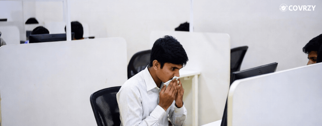 A male employee, wearing a white shirt, blowing off his nose while sit in front of a computer, around him are few vacant office chairs in white cubicles