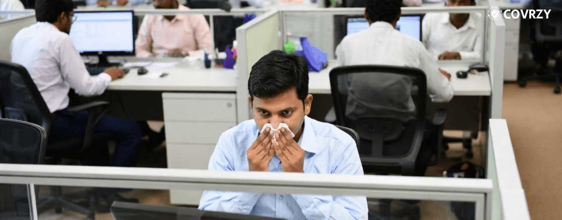 A male employee, wearing a light blue shirt, blowing off his nose while sitting in his cubicle, his surrounding has other employees working in their own cubicles