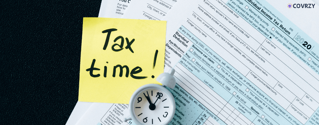 A small white clock lying on tax returns papers with a yellow sticky note saying tax time