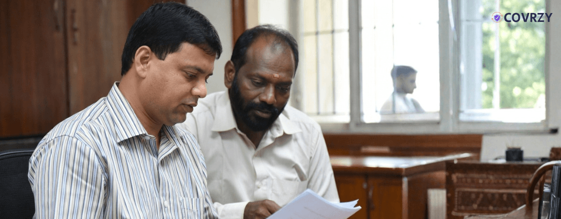 A man in a white striped shirt sitting with another man in a white shirt, both reading a document. The background is an office chamber where another person can be seen in the window behind them